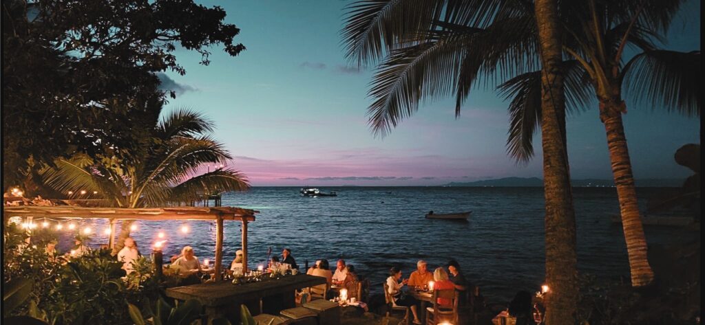 A serene seaside dining scene featuring guests enjoying a meal under string lights, with palm trees framing the view of a calm ocean and a colorful twilight sky.