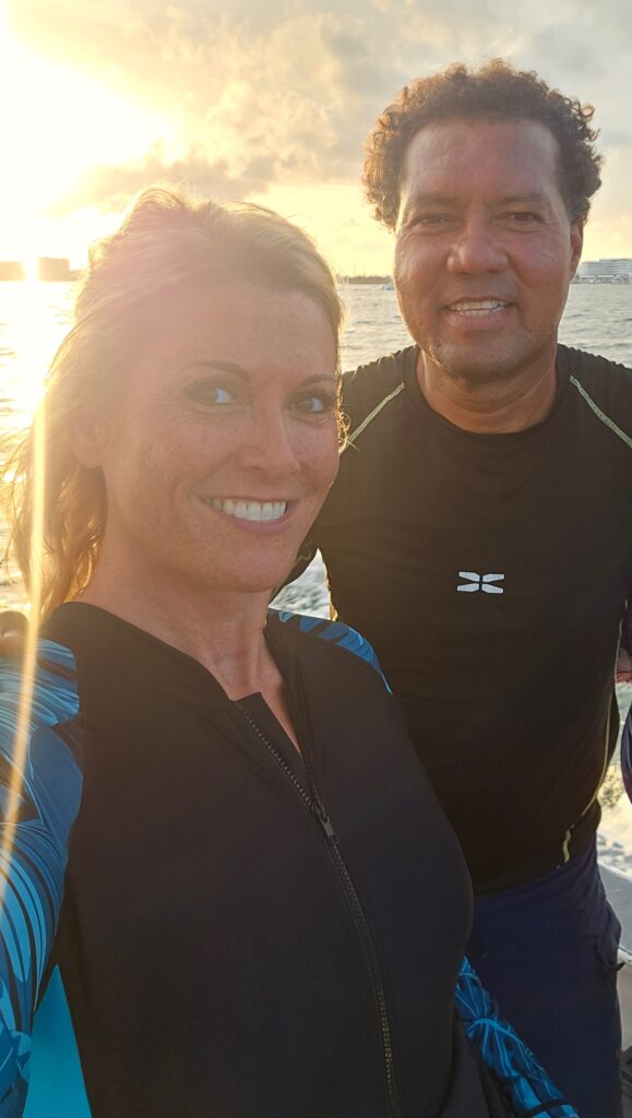 A joyful selfie of two friends at sunset, with the ocean in the background, both wearing wetsuits and smiling.