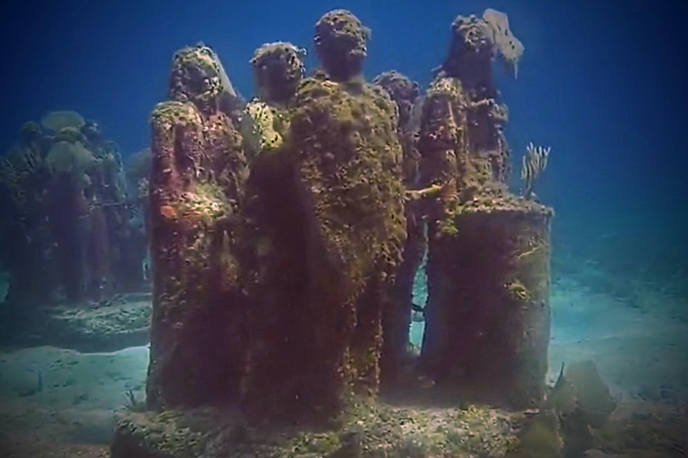 An artistic installation of submerged statues covered in marine life, set against a vibrant blue ocean backdrop.