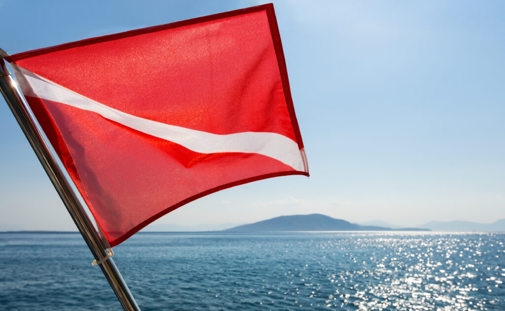 A red diving flag with a white diagonal stripe flutters over the shimmering sea and distant mountains under a clear blue sky.