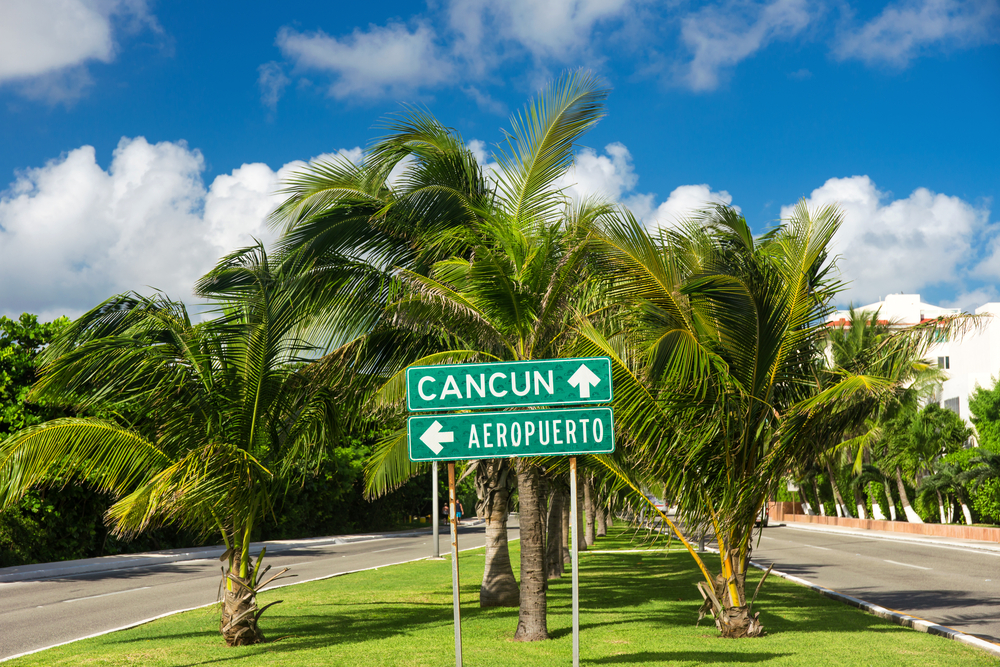 A green directional sign points towards Cancun and the airport, surrounded by palm trees under a blue sky.