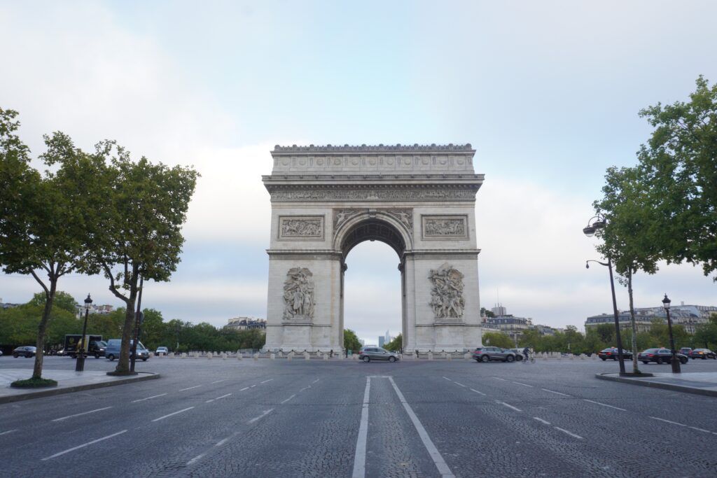 A view of the Arc de Triomphe framed by trees, with a mostly empty road leading to the monument under a cloudy sky.