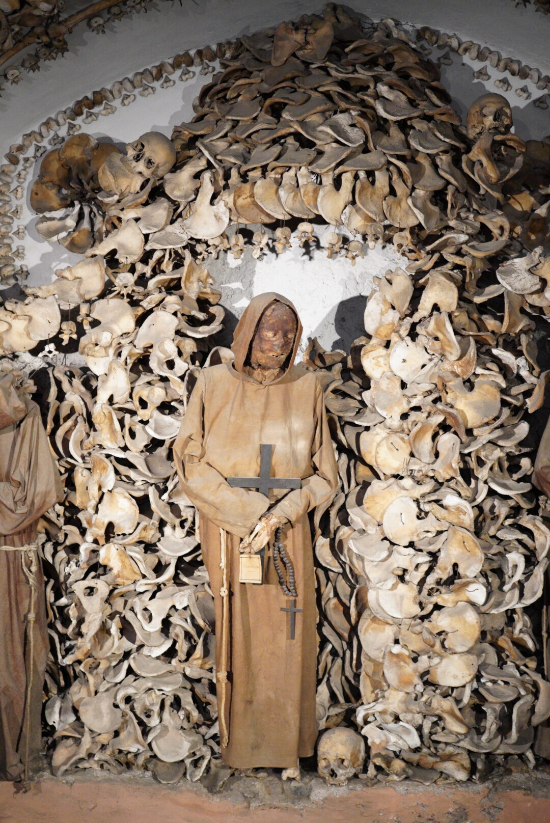 A mummified monk in a brown robe stands in front of an intricate display of human bones, including skulls and skeletal remains, forming an archway.