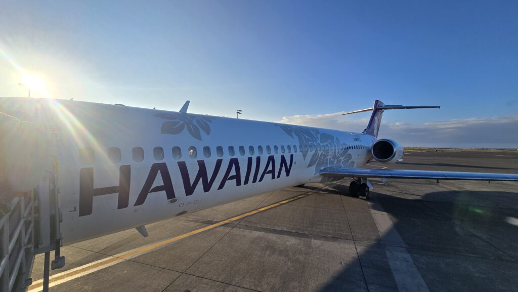 A Hawaiian Airlines airplane parked at an airport during sunset, showcasing its branding and decorative design against a clear blue sky.