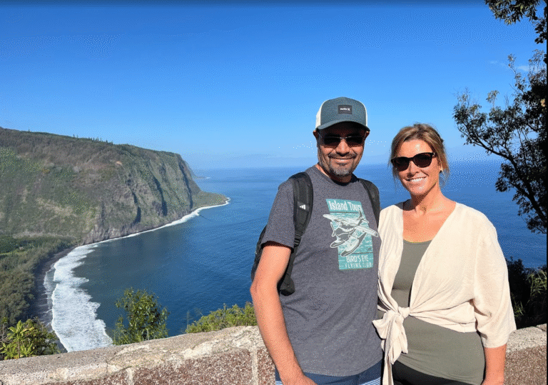 A smiling couple poses for a photo with a stunning coastal backdrop, showcasing cliffs and the ocean under a clear blue sky.