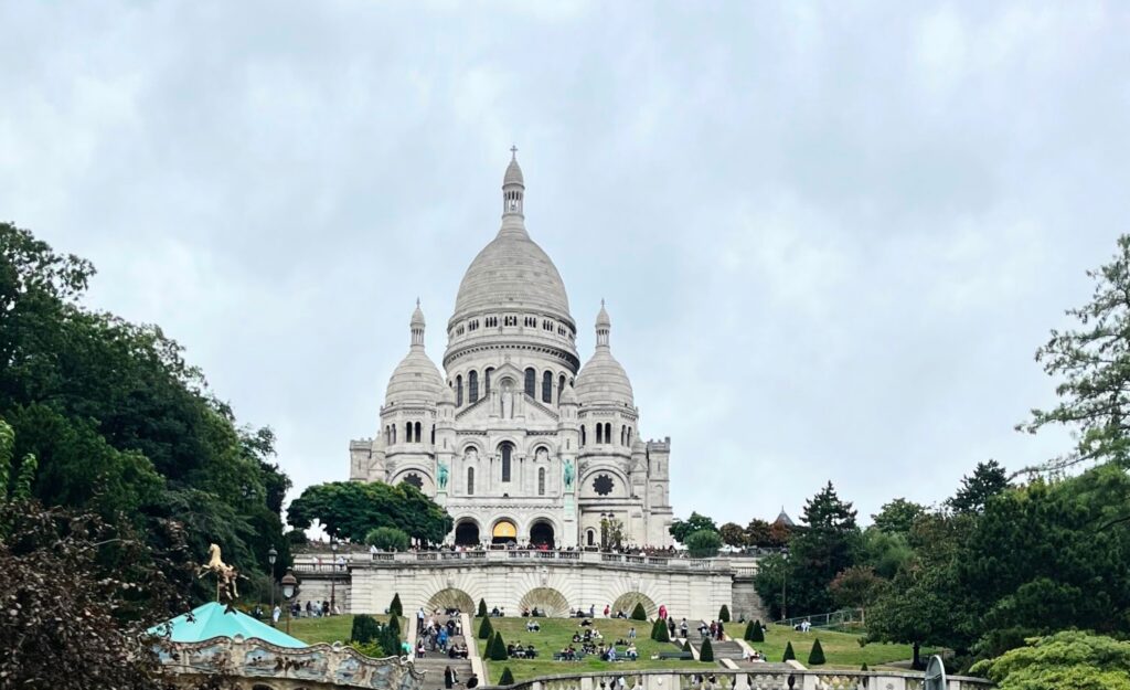 A scenic view of Sacré-Cœur Basilica surrounded by greenery and visitors, with a cloudy sky overhead.