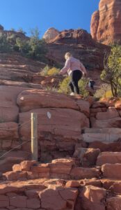 A woman climbs a steep rock path surrounded by desert vegetation and red rock formations under a clear blue sky.