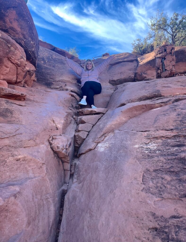 A person joyfully sitting on a rocky cliff with arms raised, surrounded by vibrant blue skies and rugged terrain.