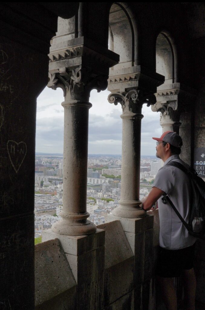 Person gazing out from a stone window at a cityscape, framed by ornate columns, under a cloudy sky.