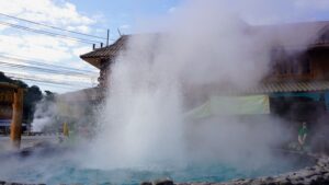 The White Temple, Chiang Rai A natural geyser erupts with steam and water near a hot springs area, surrounded by mist and a partially obscured building.