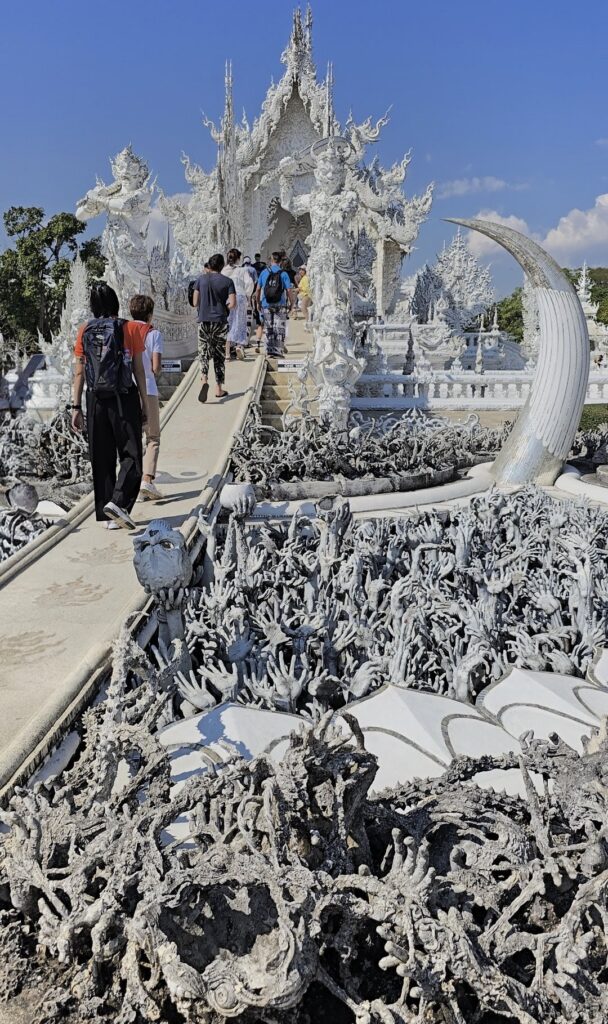 Visitors walking towards the stunning White Temple in Thailand, surrounded by intricate white sculptures and a unique landscape.