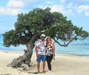 A couple poses together on a sandy beach with a distinctive tree in the background and turquoise waters.