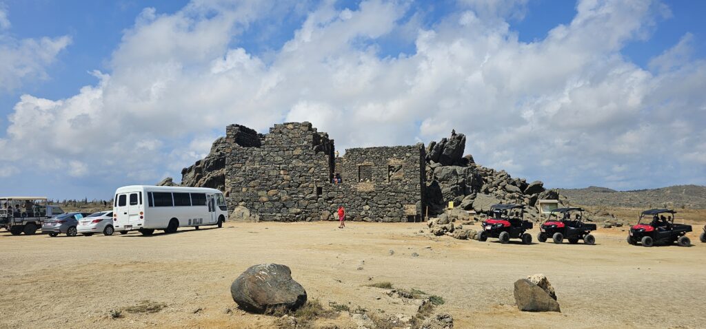 A view of stone ruins against a blue sky, surrounded by parked vehicles and rocky terrain.
