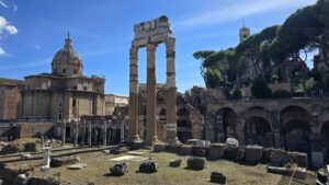 A view of ancient Roman columns among ruins, framed by modern buildings and blue skies in Rome.
