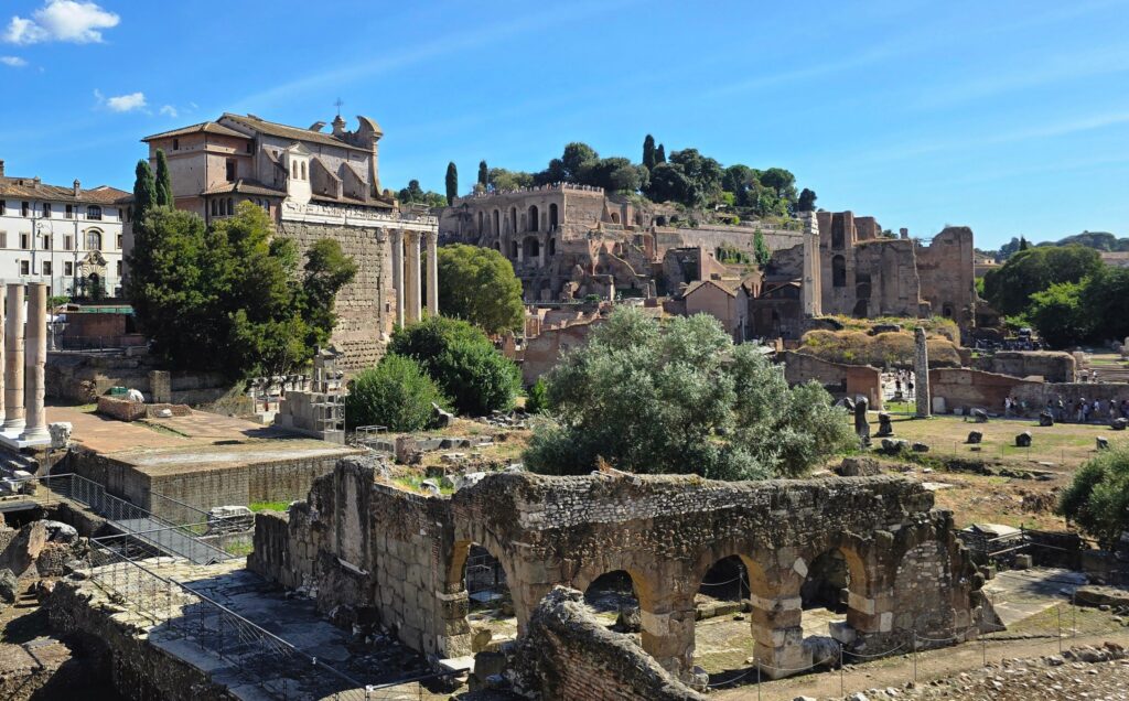 A panoramic view of the Roman Forum featuring ancient structures, lush greenery, and a clear blue sky.
