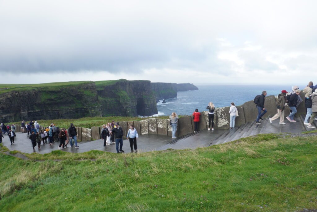 Visitors enjoying the scenic view of the Cliffs of Moher along a pathway with lush green grass and stormy skies.