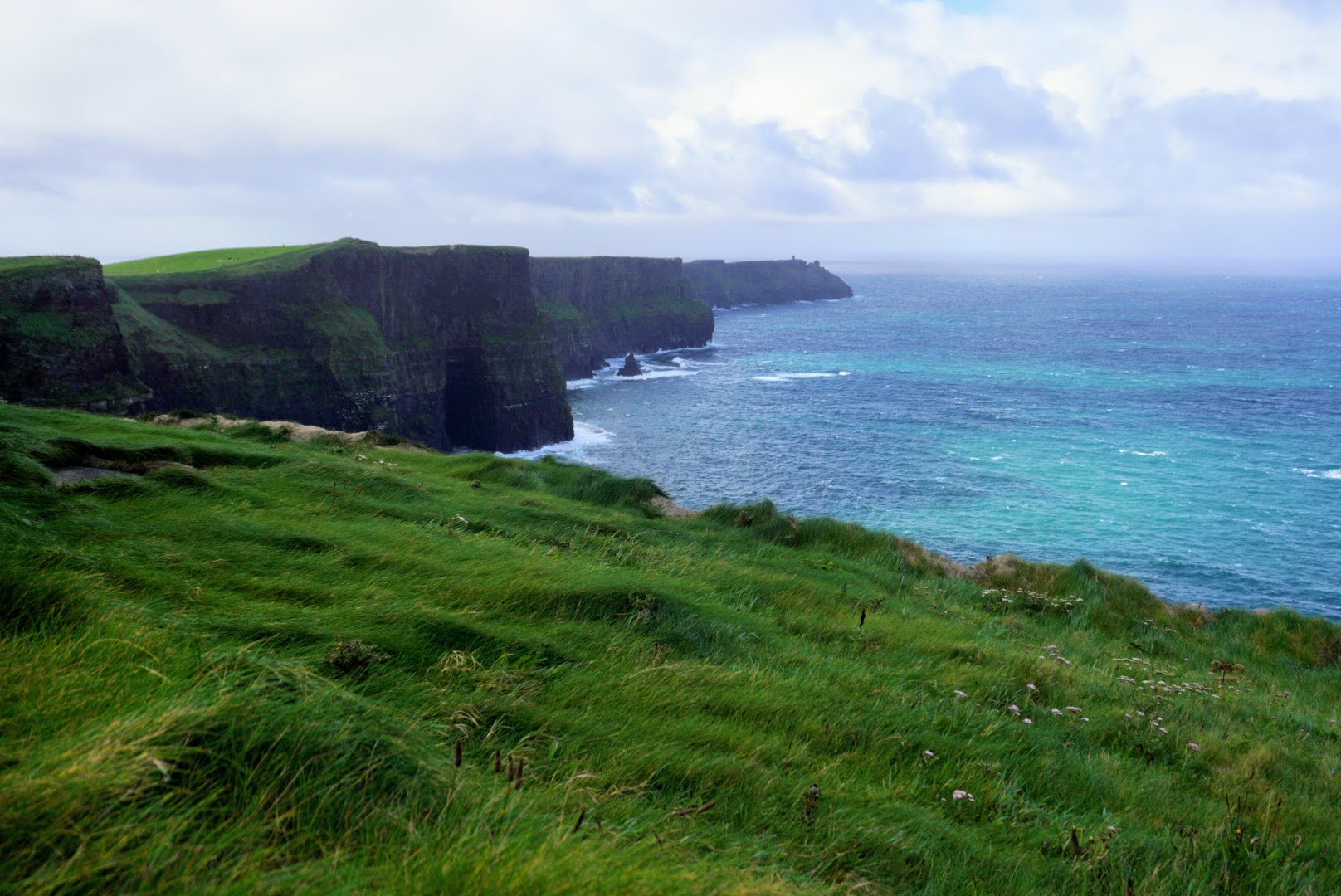 Lush green grass leads to dramatic cliffs facing a vibrant blue ocean under a cloudy sky.