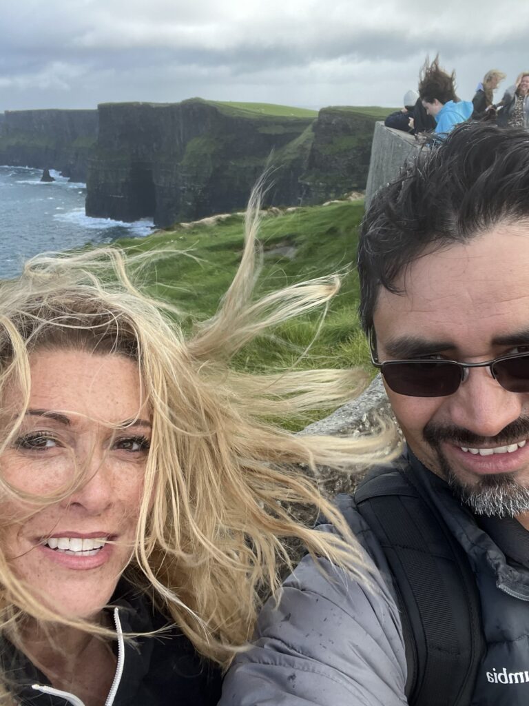 A couple enjoying a windy day at the Cliffs of Moher, with dramatic cliffs and ocean views in the background.