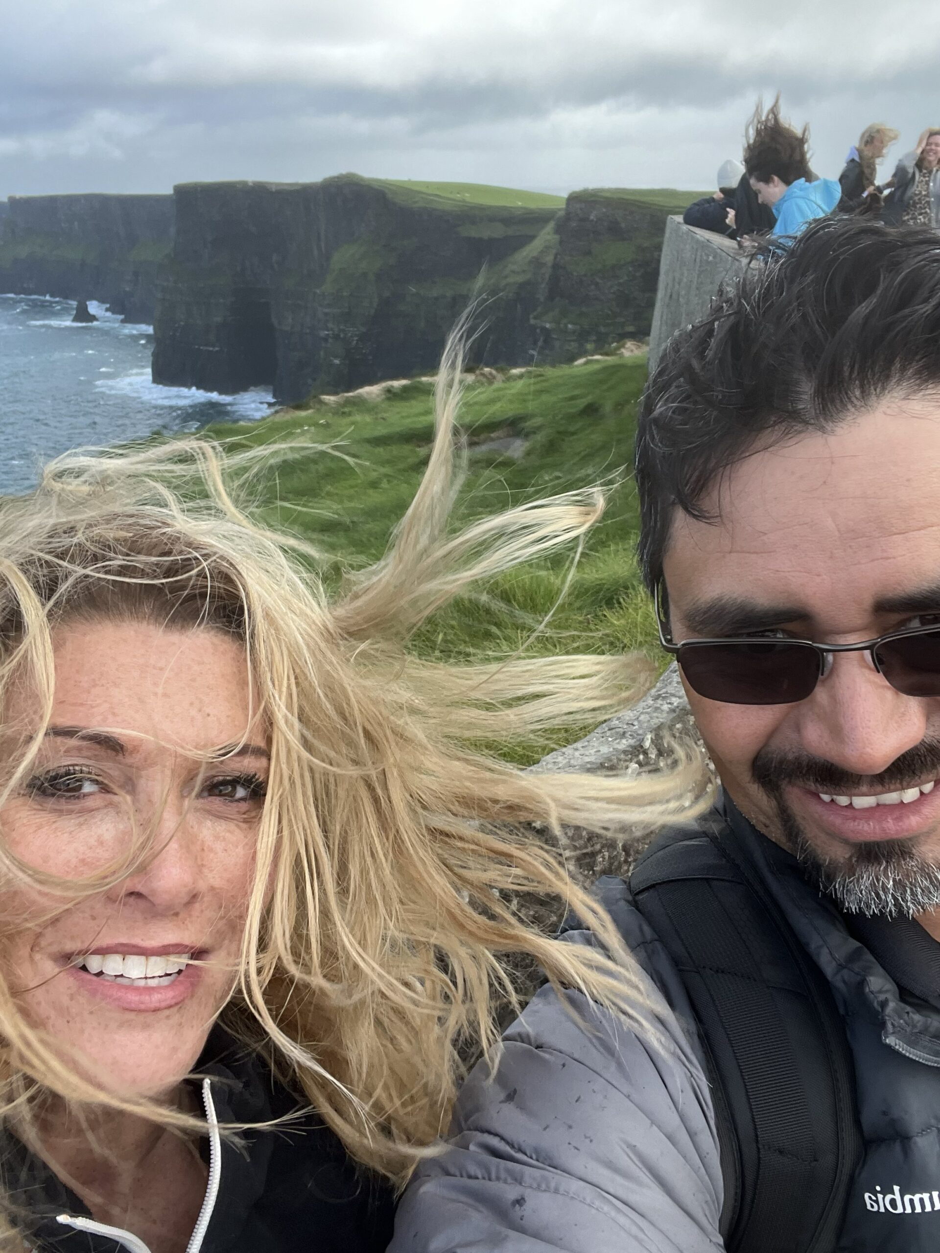 A couple enjoying a windy day at the Cliffs of Moher, with dramatic cliffs and ocean views in the background.