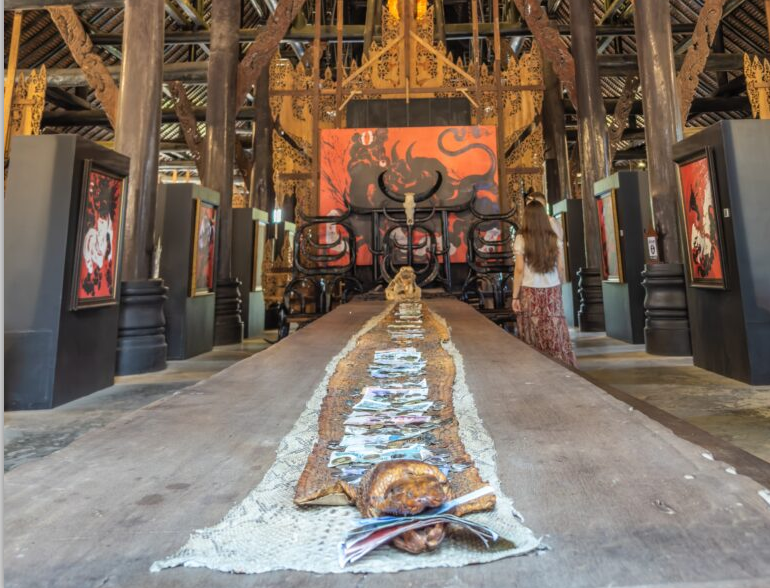 A view of a unique art installation featuring a long wooden table adorned with artifacts, while a visitor admires the vibrant artwork in a culturally rich environment.