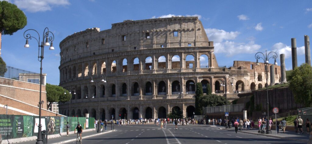 View of the ancient Colosseum with pedestrians and cyclists, surrounded by historic architecture and a blue sky.