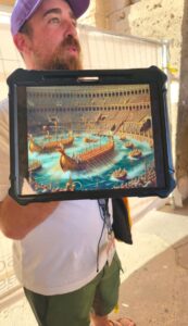 A man holds a tablet showing a vibrant illustration of ancient ships in the Colosseum, surrounded by a cheering crowd, emphasizing historical naval battles.