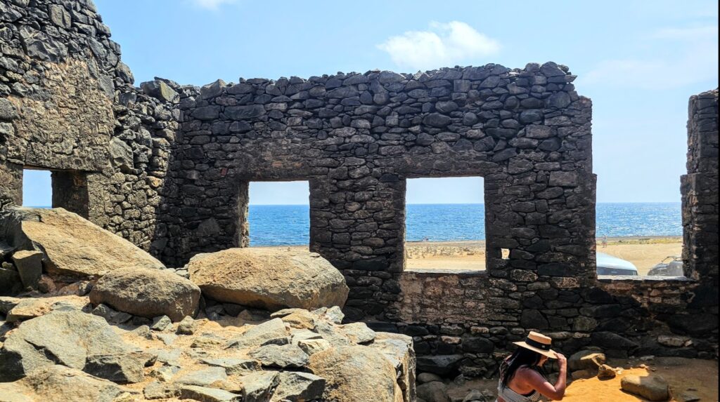 A view through stone ruins towards the ocean, with a person seated inside the structure.