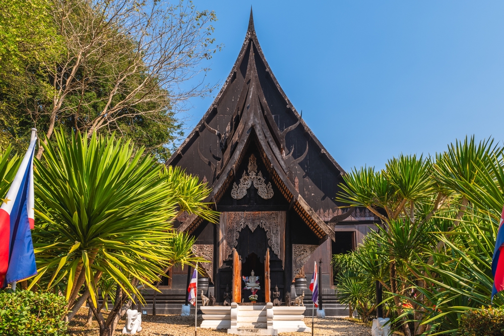 A striking black temple with intricate architectural details, surrounded by lush green vegetation and flags in the foreground.