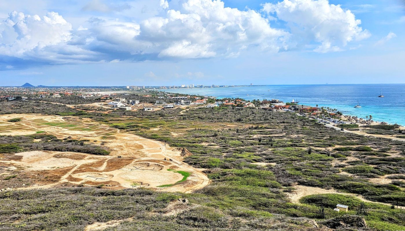 The Bushiribana Gold Mill Ruins In Aruba A panoramic view of Aruba showcasing a mix of green vegetation, desert terrain, and the blue ocean in the background, with residential areas lining the coast.