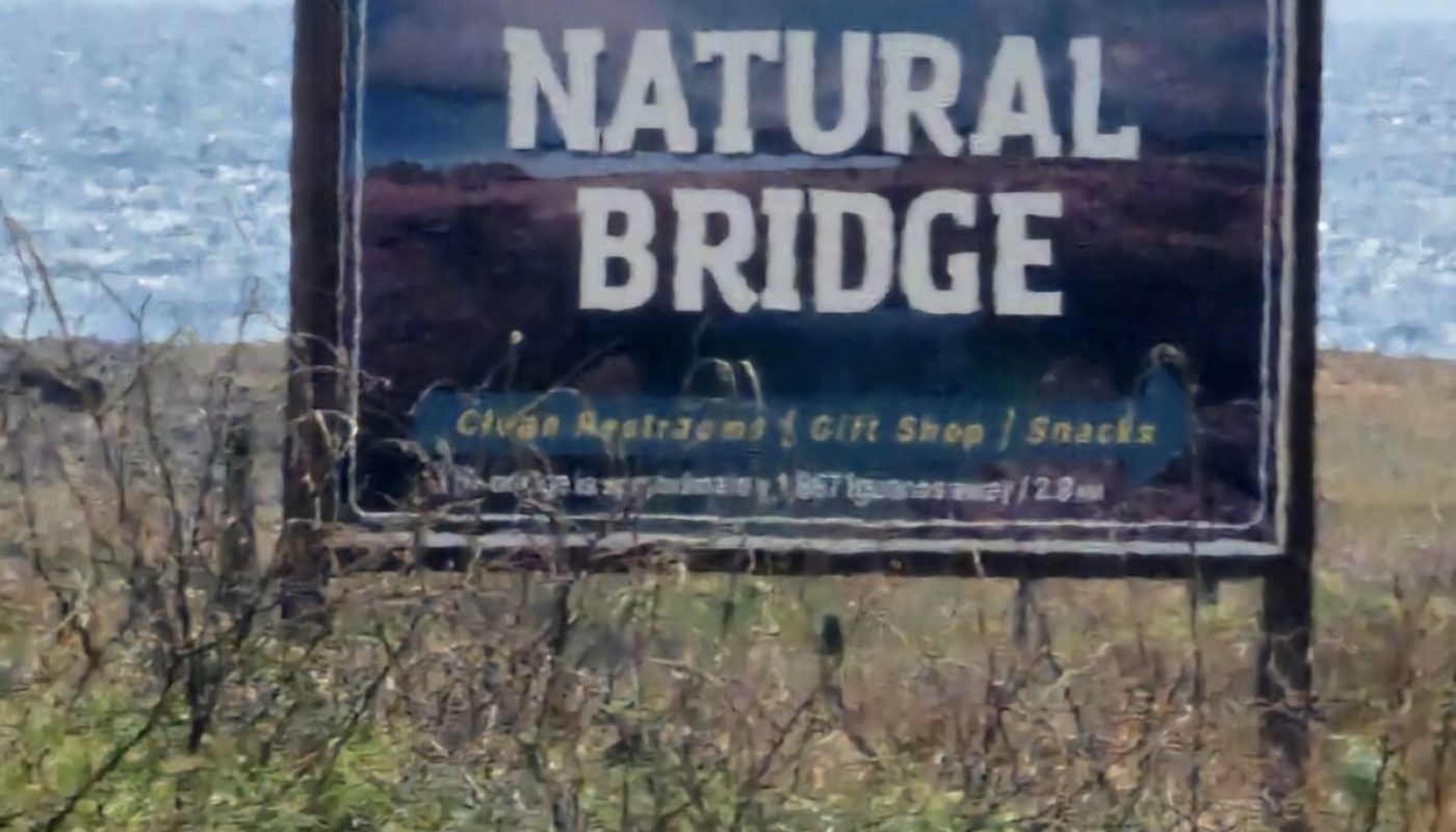 The Bushiribana Gold Mill Ruins In Aruba A sign for Natural Bridge, located near a body of water, with information about dining, shopping, and snacks.