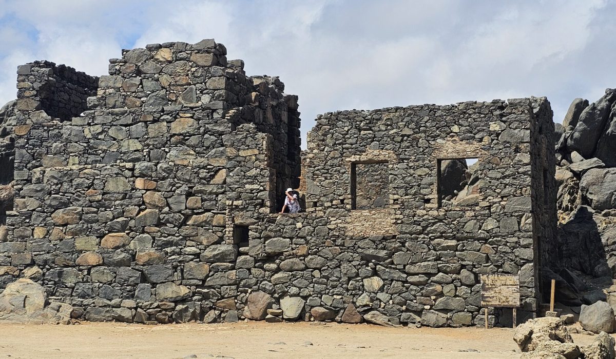 The Bushiribana Gold Mill Ruins In Aruba A weathered stone building with missing windows, partially hidden by rocks, and two people exploring the site under a cloudy sky.