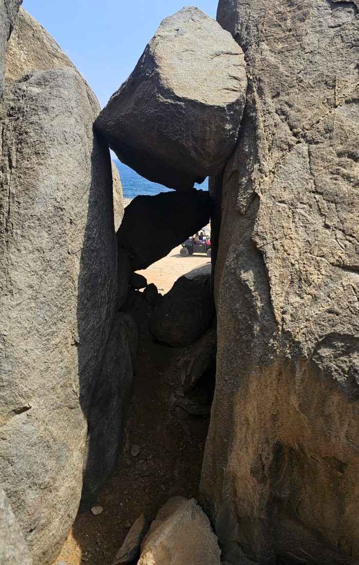 The Bushiribana Gold Mill Ruins In Aruba A narrow gap between large boulders reveals a scenic coastal landscape beyond.