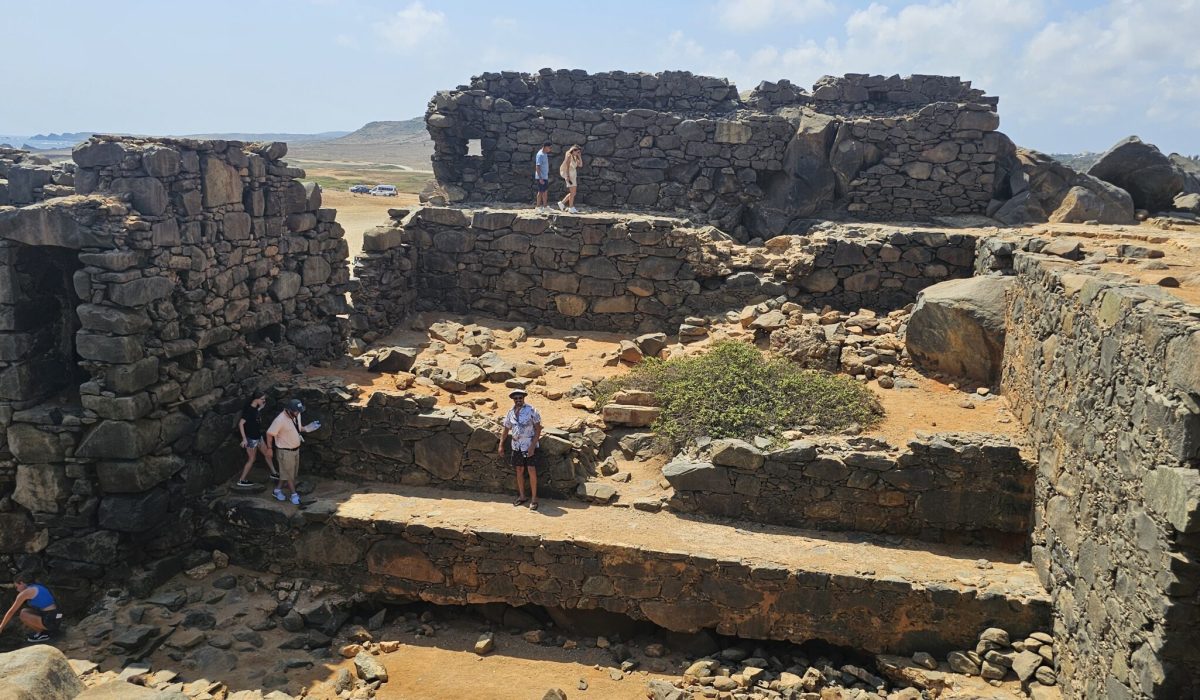 The Bushiribana Gold Mill Ruins In Aruba A view of stone ruins with tourists exploring the area, featuring uneven surfaces and surrounding landscapes.