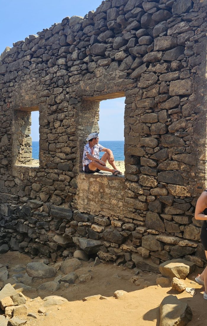 The Bushiribana Gold Mill Ruins In Aruba A person sitting in a window of a stone ruin, with ocean views and sandy ground in the foreground.