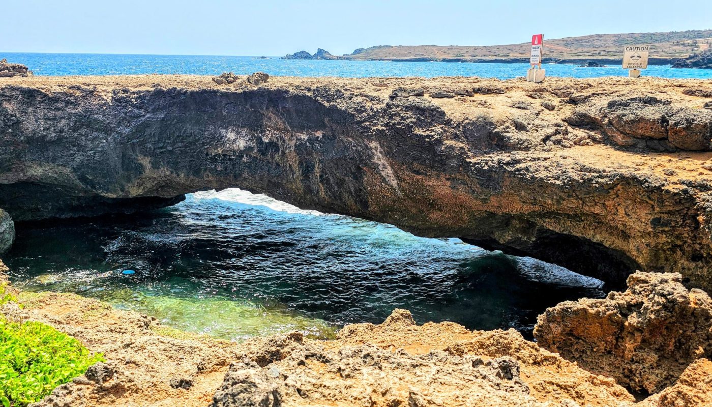The Bushiribana Gold Mill Ruins In Aruba A scenic view of a natural rock arch overlooking the ocean, with caution signs visible on the shore.