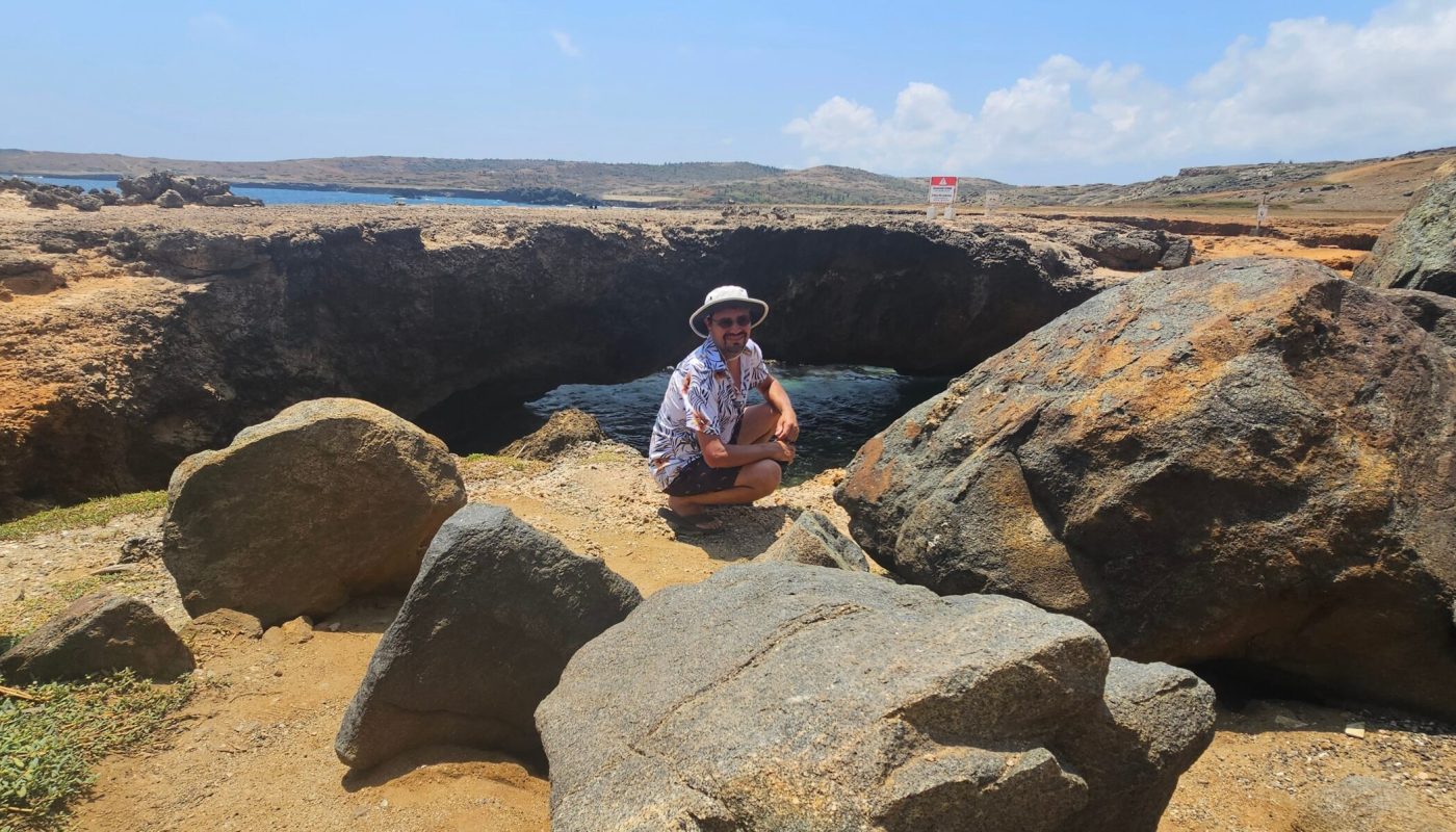 The Bushiribana Gold Mill Ruins In Aruba A person in a floral shirt and white hat crouches near large rocks by the sea, with a caution sign visible in the background.