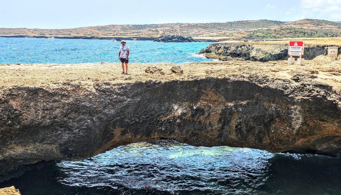 The Bushiribana Gold Mill Ruins In Aruba A person standing on a rocky ledge overlooking the ocean, with a natural archway formed in the rocks and warning signs in the background.