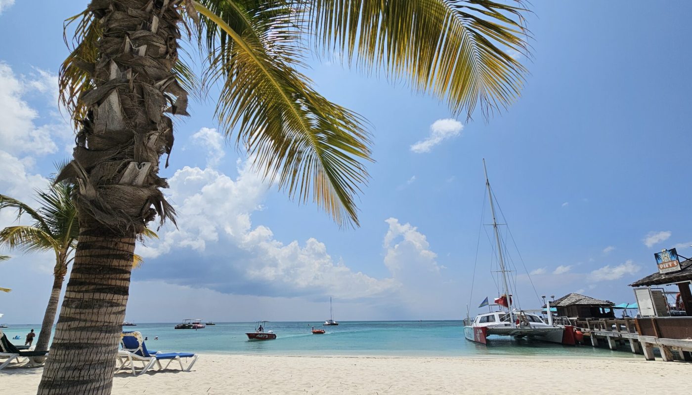 The Bushiribana Gold Mill Ruins In Aruba A serene beach scene featuring palm trees, clear blue water, and boats anchored in the distance under a bright sky with fluffy clouds.
