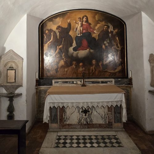 A serene chapel featuring an altar adorned with a detailed religious painting of the Madonna and Child, surrounded by figures, set against an arched wall.