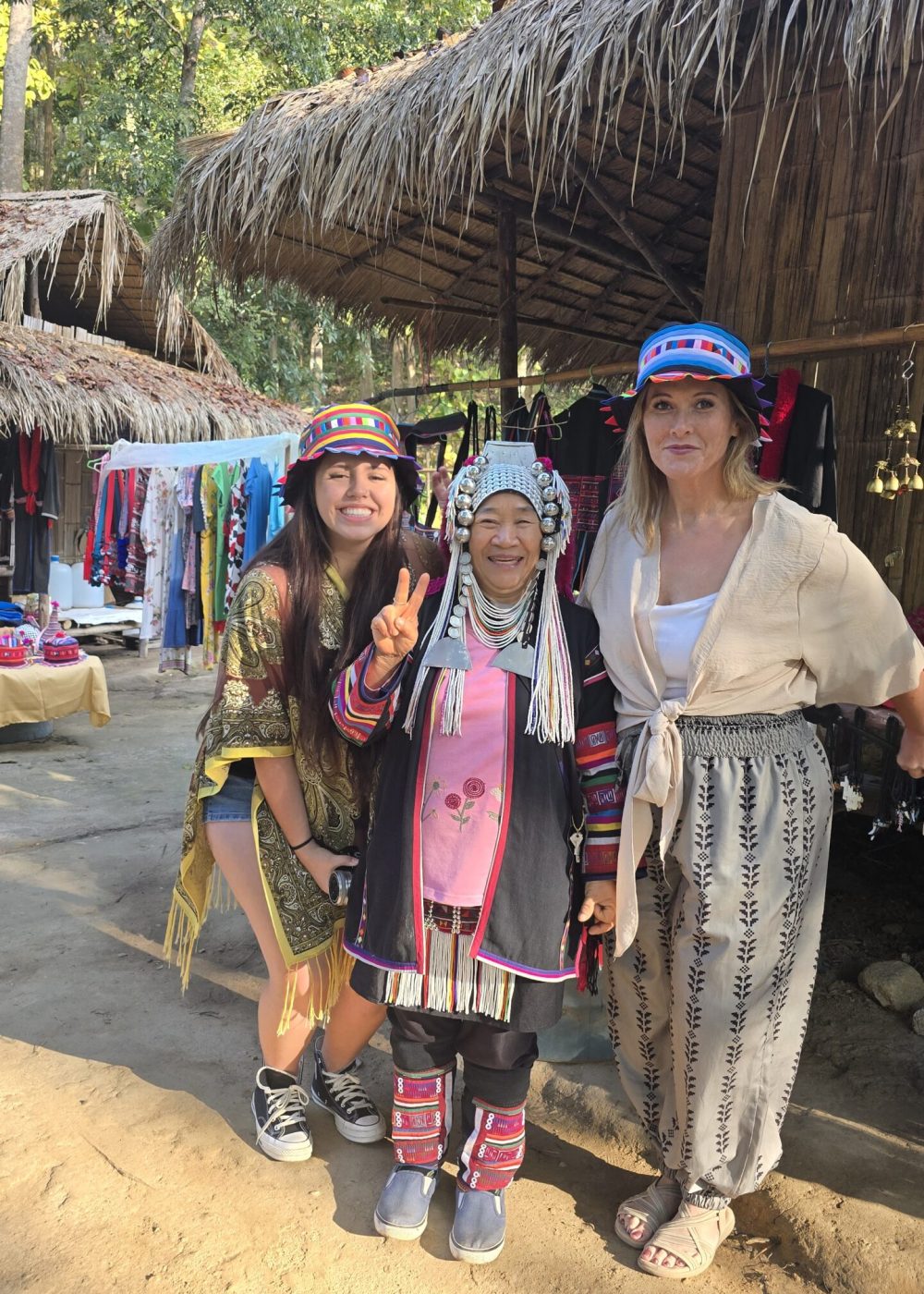 The White Temple, Chiang Rai Three women posing together in colorful traditional attire, surrounded by huts and market stalls in a lush forest setting.