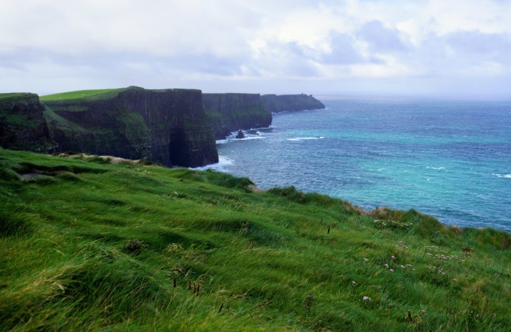 Lush green grass leads to dramatic cliffs facing a vibrant blue ocean under a cloudy sky.