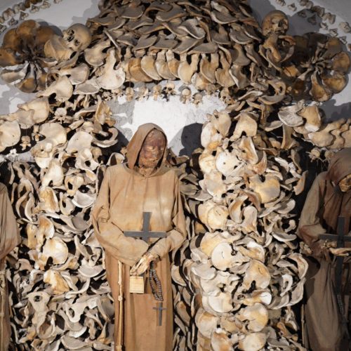 A chilling arrangement of skeletons and skulls in a chapel, featuring mummified figures in hooded robes holding religious icons, surrounded by a wall of bones.