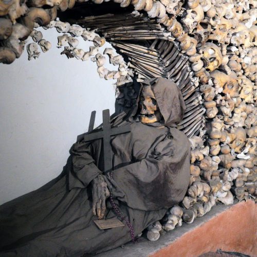 A cloaked skeleton holding a cross is seated against a backdrop of intricately arranged human skulls and bones, creating an eerie atmosphere.