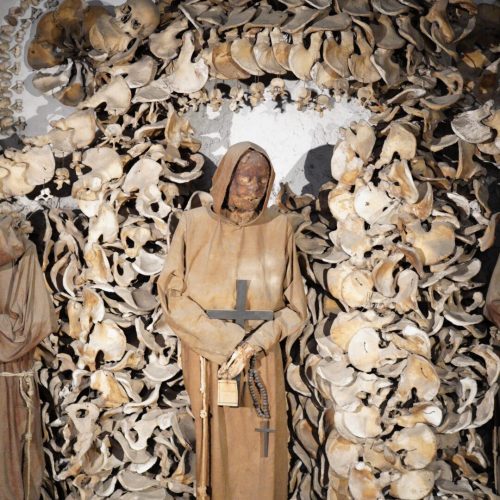 A mummified monk in a brown robe stands in front of an intricate display of human bones, including skulls and skeletal remains, forming an archway.