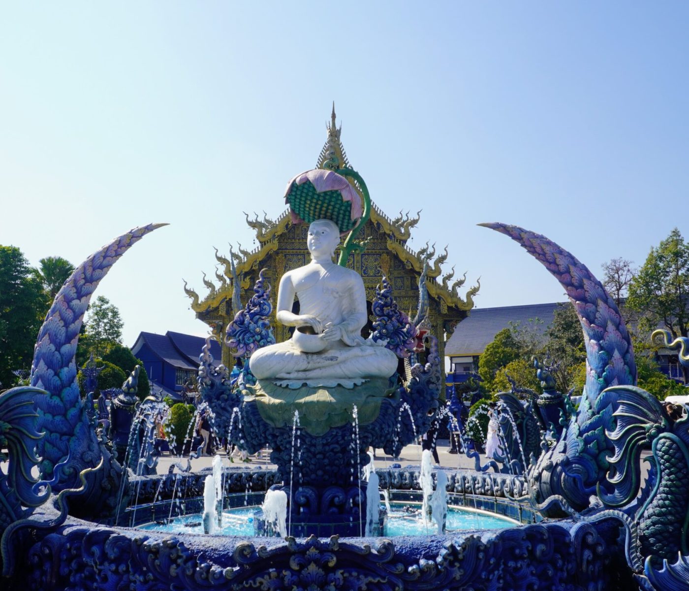 A stunning Buddha statue is centrally featured, surrounded by ornate blue and purple sculptures with a backdrop of a decorative building against a clear blue sky.