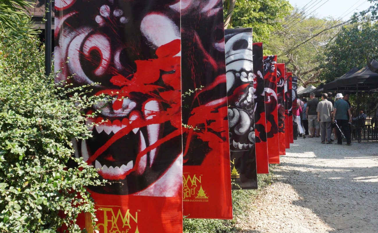 Baan Dam: The Black House Museum Colorful banners featuring stylized animal heads line a pathway, surrounded by greenery and visitors engaging with the art.
