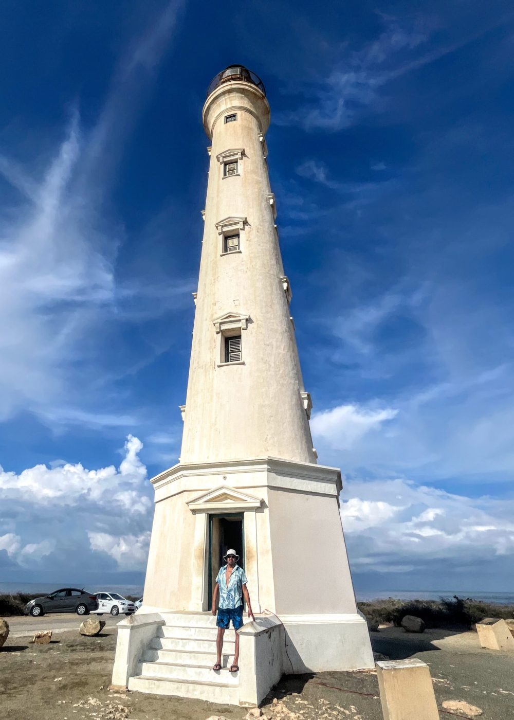 The Bushiribana Gold Mill Ruins In Aruba A person stands at the base of a tall lighthouse with a bright blue sky and clouds in the background.