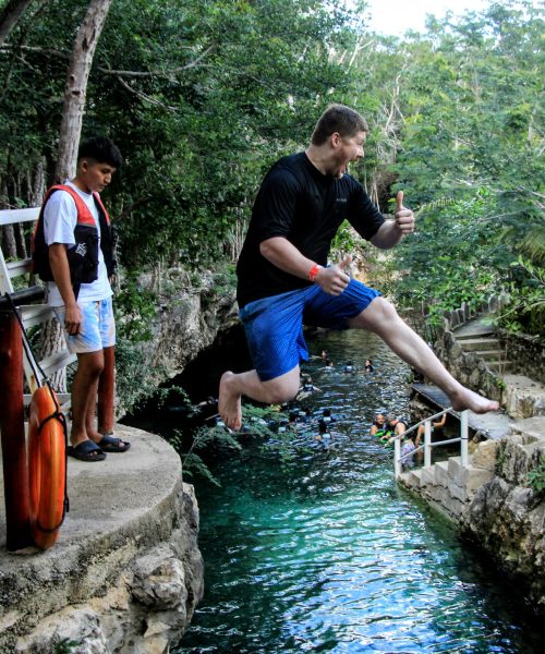 Man jumping enthusiastically into a cenote while giving a thumbs-up, with others swimming in the clear blue water below