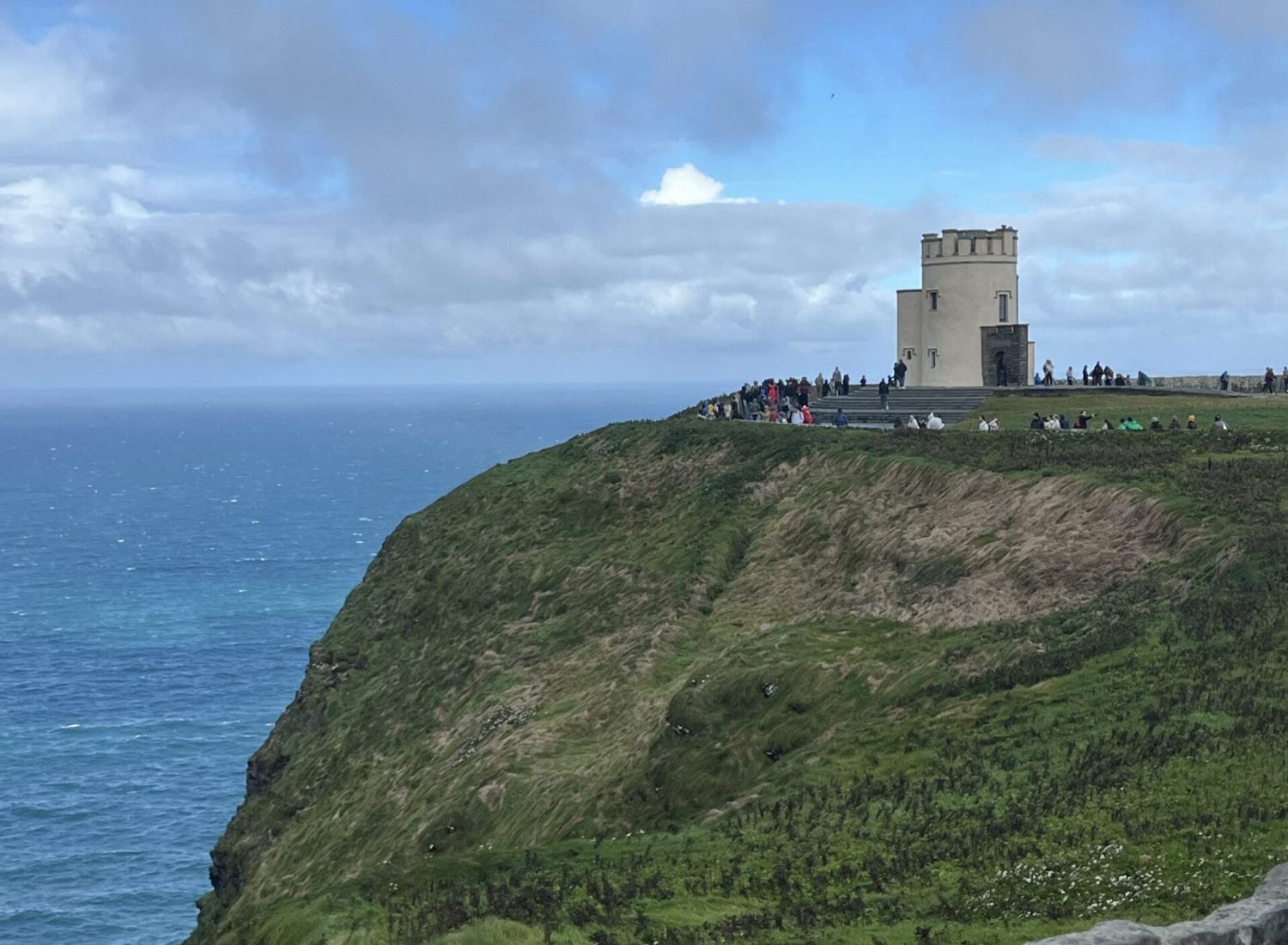 A scenic view of a green cliff featuring a historic tower, with a crowd of visitors admiring the ocean and dramatic sky in the background.