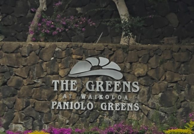 A stone wall sign displaying "The Greens at Waikoloa Paniolo Greens" surrounded by colorful flowers and greenery.